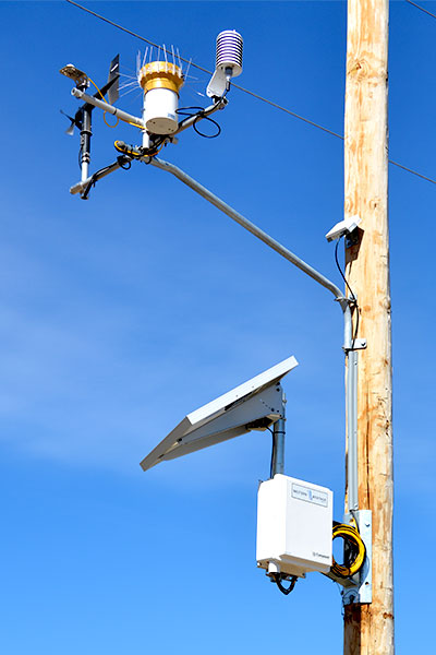 Close-up of a satellite-enabled, pole-mounted weather monitoring system with wind, temperature, and precipitation sensors, solar panel, and data logger unit against a clear sky